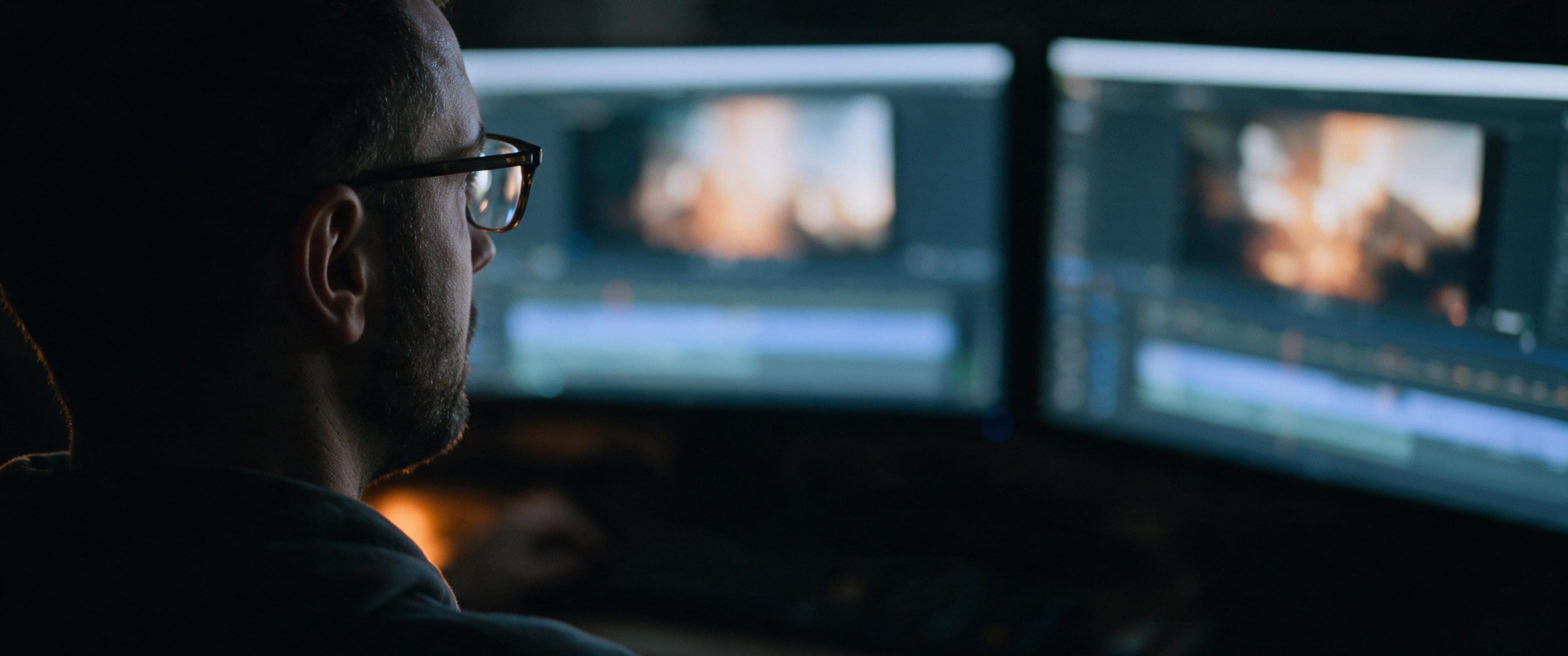 Joshua Davies at an edit, seen from behind in a dark room, his face lit only by two screens holding a graded shot and a timeline.